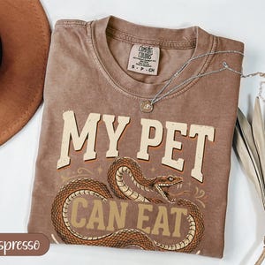 Puede incluir: Una camiseta gris carbón de manga corta con el gráfico de una serpiente marrón enroscada. El texto MY PET CAN EAT YOUR PET aparece en una fuente vintage llamativa. La prenda tiene un aspecto desgastado y lavado con cuello redondo.