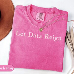 Könnte beinhalten: Ein leuchtend rosa Kurzarm-T-Shirt mit Rundhalsausschnitt und dem in Weiß auf die Brust gedruckten Text Let Data Reign. Das Shirt hat ein Comfort Colors-Etikett am Innenkragen und wird mit einer goldfarbenen Halskette mit kleinem Anhänger kombiniert.