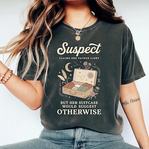 May include: Dark gray t-shirt with the word "Suspect" in a vintage font. The shirt features a suitcase graphic with the text "Claims she packed light, but her suitcase would suggest otherwise."