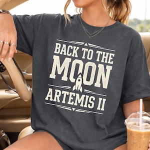 Op de afbeelding: Donkergrijs T-shirt met de witte tekst "BACK TO THE MOON ARTEMIS II". Een witte raket-afbeelding staat onder "MOON". Het shirt heeft een relaxte pasvorm.