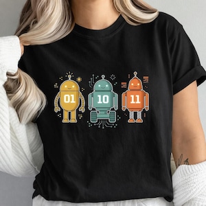 Op de afbeelding: Zwart t-shirt met een illustratie van drie kleurrijke robots. De robots zijn geel, blauwgroen en oranje, elk met een uniek ontwerp en de binaire getallen 01, 10 en 11 op hun borst. Het ontwerp is in cartoonstijl.
