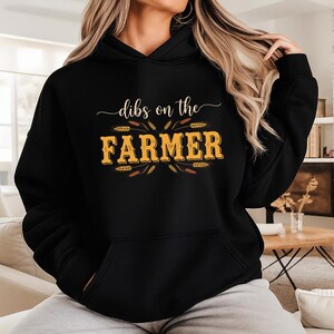 May include: Black hoodie with the text "dibs on the FARMER" in a gold and brown design. The hoodie has a front pocket and a drawstring hood. The text is surrounded by wheat stalks.