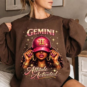 May include: A brown sweatshirt with a graphic of a woman wearing a pink cap with the Gemini symbol. The words "GEMINI" and "Attitude Activated" are displayed in a decorative font. The design includes gold accents and star details.