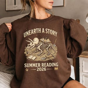 May include: Brown sweatshirt with the text "UNEARTH A STORY" above a graphic of a dinosaur skeleton with books and tools. The text "SUMMER READING - 2026" is below the graphic. The sweatshirt is a solid brown color.