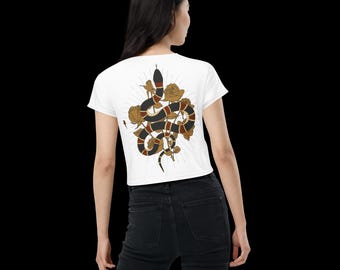 Puede incluir: Top corto blanco con un gráfico detallado de una serpiente y rosas. La serpiente es negra, marrón y beige, con rosas doradas y un diseño de rayos de sol. La parte superior es de manga corta y de estilo casual.