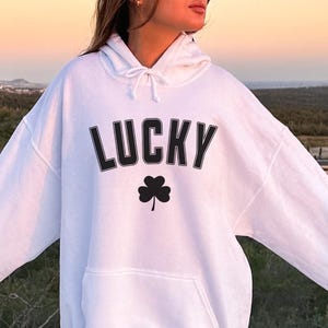 Puede incluir: Sudadera blanca con la palabra "LUCKY" en letras negras y un trébol negro. La sudadera tiene un bolsillo delantero y una capucha con cordón. Las mangas son largas y el ajuste es holgado.