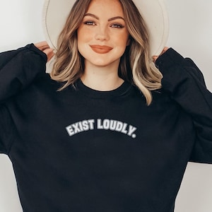 May include: Black sweatshirt with the text "EXIST LOUDLY" in white, block letters. The sweatshirt has long sleeves and a crew neck. The model is wearing a cream-colored hat.