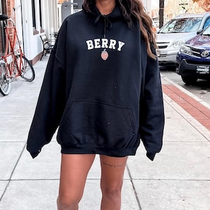May include: Black hooded sweatshirt with the word "BERRY" in white block letters and a small strawberry graphic. The sweatshirt has a front pocket and long sleeves. The person is wearing white sneakers.