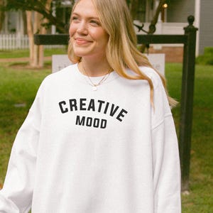 以下が含まれることがあります： 黒字で「CREATIVE MOOD」とプリントされた白いスウェットシャツ。 スウェットシャツはクルーネックで、柔らかい素材でできているようです。 画像は晴れた日に屋外で撮影されました。