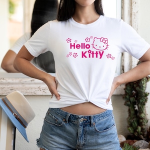 May include: White t-shirt with a pink Hello Kitty design and floral details. The top is knotted at the waist, worn with denim shorts. A blue and beige hat is visible.