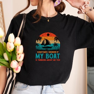 May include: Black t-shirt featuring a sunset design with a sailboat, palm trees, and the text "Sometimes I wonder if my boat is thinking about me too." The graphic is in shades of orange, blue, and green.