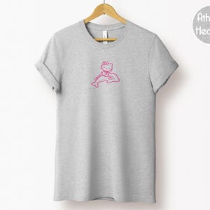 May include: A heather gray t-shirt with a pink graphic of a cartoon cat riding a dolphin. The t-shirt is hanging on a wooden hanger against a white background. The text "Athletic Heather" is in a circle on the upper right.
