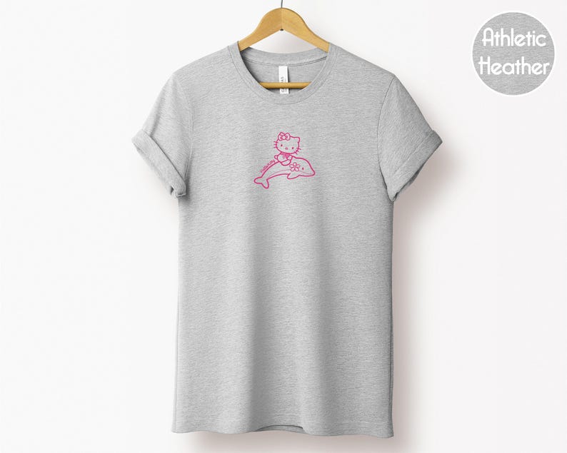 May include: A heather gray t-shirt with a pink graphic of a cartoon cat riding a dolphin. The t-shirt is hanging on a wooden hanger against a white background. The text "Athletic Heather" is in a circle on the upper right.