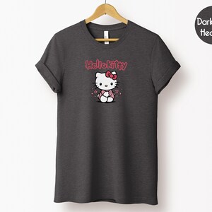 May include: Dark grey heather t-shirt featuring a Hello Kitty graphic. The graphic includes the text "Hello Kitty" in pink above an illustration of Hello Kitty wearing a pink bow and dress. The t-shirt is hanging on a wooden hanger.