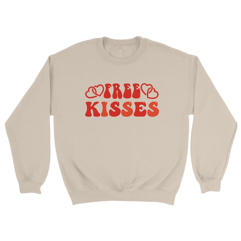 May include: A beige sweatshirt featuring the text "FREE KISSES" in a retro red and orange font, with heart motifs. This casual sweatshirt is designed for comfort.