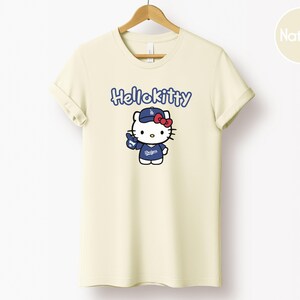 May include: Cream-coloured t-shirt with the blue text "Hello Kitty" above a cartoon Hello Kitty character wearing a blue baseball cap and uniform with the Dodgers logo. The shirt is hanging on a wooden hanger.
