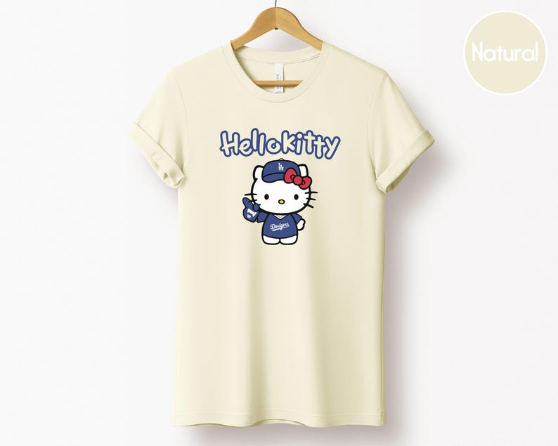 May include: Cream-coloured t-shirt with the blue text "Hello Kitty" above a cartoon Hello Kitty character wearing a blue baseball cap and uniform with the Dodgers logo. The shirt is hanging on a wooden hanger.