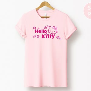 May include: A pale pink t-shirt featuring the words "Hello Kitty" and a fuchsia graphic of Hello Kitty. The t-shirt is displayed on a wooden hanger against a white backdrop. The sleeves are rolled up.