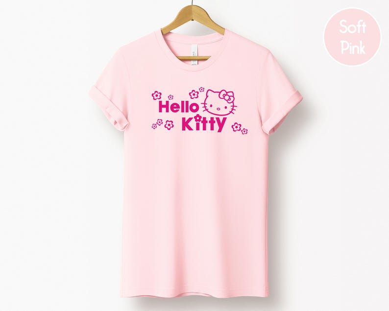 May include: A pale pink t-shirt featuring the words "Hello Kitty" and a fuchsia graphic of Hello Kitty. The t-shirt is displayed on a wooden hanger against a white backdrop. The sleeves are rolled up.