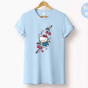Op de afbeelding: Lichtblauw T-shirt met een cartoonfiguur met een rode strik, blauwe overall en gele sjaal, naast een tak met rode kersen en witte bloemen. Het T-shirt hangt aan een houten kleerhanger.