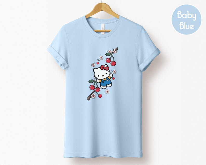 Op de afbeelding: Lichtblauw T-shirt met een cartoonfiguur met een rode strik, blauwe overall en gele sjaal, naast een tak met rode kersen en witte bloemen. Het T-shirt hangt aan een houten kleerhanger.