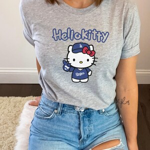May include: A light grey t-shirt with the blue text "Hellokitty". The shirt features a graphic of Hello Kitty wearing a Dodgers baseball cap and uniform, with a red bow and a blue "1" finger pointing up.
