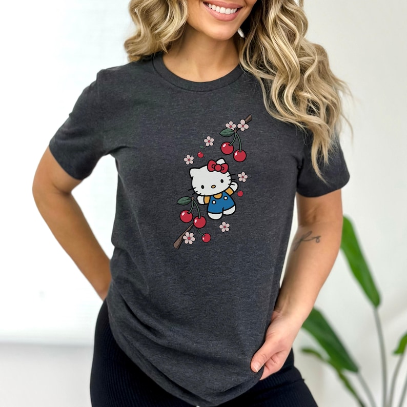 Op de afbeelding: Donkergrijs T-shirt met een cartoonfiguur met een rode strik en blauwe overall, met een kersentakontwerp. De kersen zijn rood en de bloemen roze en wit.