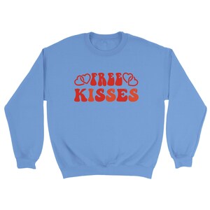 May include: A pale blue sweatshirt featuring the text "FREE KISSES" in a retro red and orange font, with heart motifs. The sweatshirt has a crew neck and long sleeves.