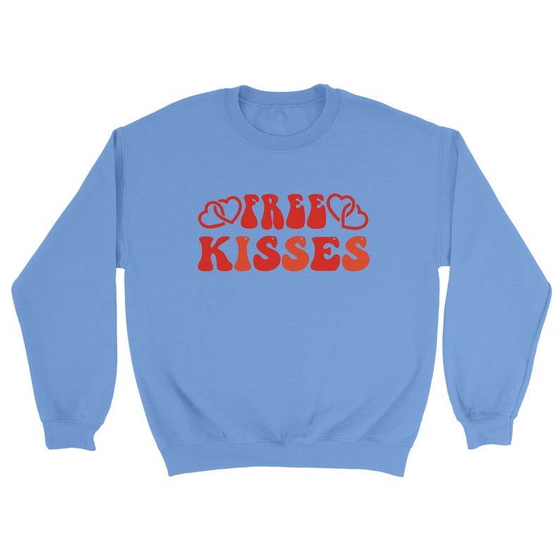 May include: A pale blue sweatshirt featuring the text "FREE KISSES" in a retro red and orange font, with heart motifs. The sweatshirt has a crew neck and long sleeves.
