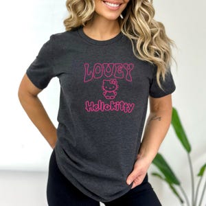 Op de afbeelding: Donkergrijs t-shirt met de roze tekst "LOVEY" boven een Hello Kitty-afbeelding en het woord "Hellokitty". Het t-shirt heeft een ronde hals.