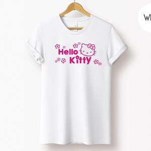 May include: White t-shirt featuring a pink Hello Kitty graphic and the words "Hello Kitty". The design includes flowers around the text and character. The shirt is displayed on a wooden hanger.