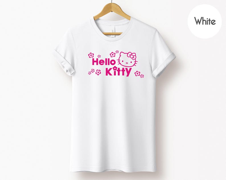 May include: White t-shirt featuring a pink Hello Kitty graphic and the words "Hello Kitty". The design includes flowers around the text and character. The shirt is displayed on a wooden hanger.