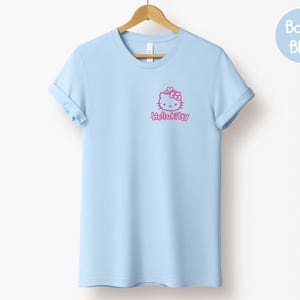 May include: A baby blue t-shirt with rolled-up sleeves, hanging on a wooden hanger. The shirt features a pink Hello Kitty graphic on the front. A light blue circle with the words "Baby Blue" is in the upper right corner.