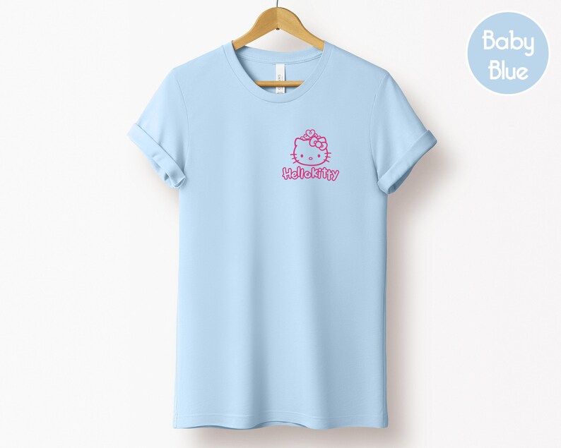 May include: A baby blue t-shirt with rolled-up sleeves, hanging on a wooden hanger. The shirt features a pink Hello Kitty graphic on the front. A light blue circle with the words "Baby Blue" is in the upper right corner.