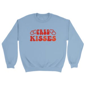 May include: A pale blue sweatshirt featuring the text "FREE KISSES" in a retro red and orange font, with heart motifs. The top has a crew neck and long sleeves.
