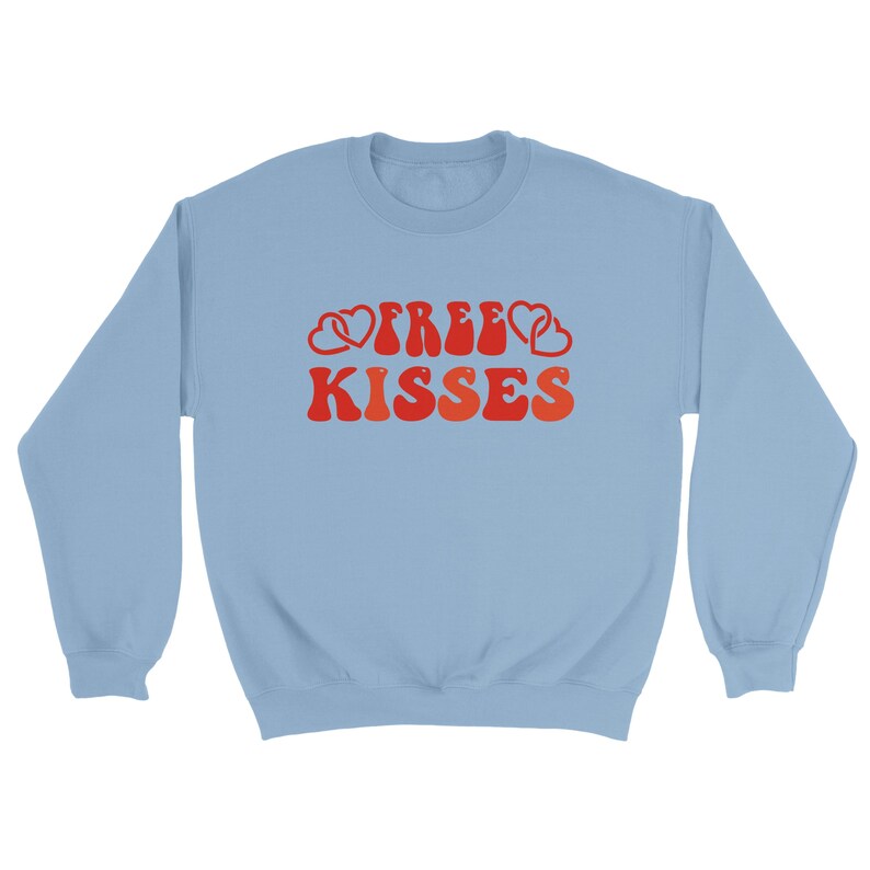 May include: A pale blue sweatshirt featuring the text "FREE KISSES" in a retro red and orange font, with heart motifs. The top has a crew neck and long sleeves.