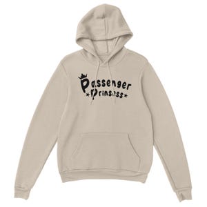 May include: A beige hooded sweatshirt with a front pocket. The front features the black text "Passenger Prinssss", with a crown above "Passenger" and stars around "Prinssss".
