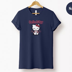 May include: Navy blue t-shirt featuring a Hello Kitty graphic. The graphic shows Hello Kitty wearing a pink dress and bow. The text "Hello Kitty" is above the character in pink. The t-shirt is hanging on a wooden hanger.