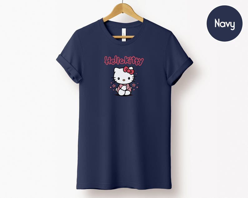 May include: Navy blue t-shirt featuring a Hello Kitty graphic. The graphic shows Hello Kitty wearing a pink dress and bow. The text "Hello Kitty" is above the character in pink. The t-shirt is hanging on a wooden hanger.