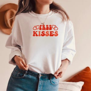 Può includere: Felpa bianca con la scritta "FREE KISSES" in rosso, con un design in stile retrò. Il disegno include forme di cuore. La felpa è a maniche lunghe e ha uno scollo tondo.