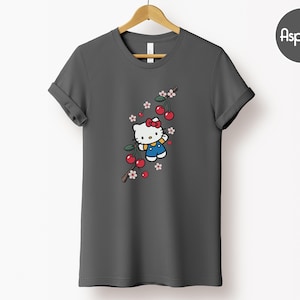 Op de afbeelding: Donkergrijs t-shirt met een cartoon illustratie van Hello Kitty met een rode strik en blauwe overall, omringd door rode kersen en roze-witte bloemen. Het ontwerp is op een tak.