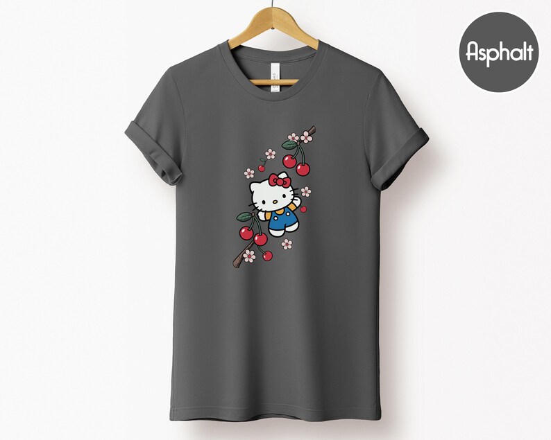 Op de afbeelding: Donkergrijs t-shirt met een cartoon illustratie van Hello Kitty met een rode strik en blauwe overall, omringd door rode kersen en roze-witte bloemen. Het ontwerp is op een tak.