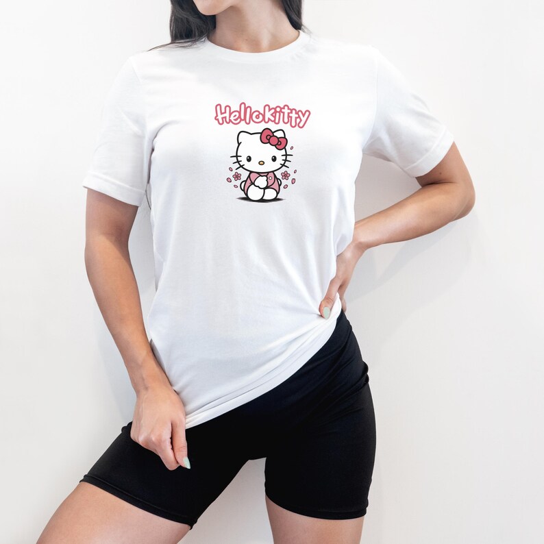 May include: White t-shirt featuring a Hello Kitty graphic. The graphic includes the text "Hello Kitty" in pink above an illustration of the character. The shirt is short-sleeved and appears to be made of a soft material.