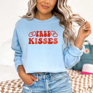 May include: A pale blue sweatshirt featuring the text "FREE KISSES" in red, with heart motifs. The design has a retro aesthetic. A casual top.