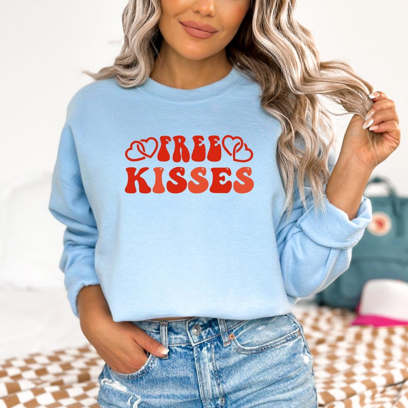 May include: A pale blue sweatshirt featuring the text "FREE KISSES" in red, with heart motifs. The design has a retro aesthetic. A casual top.