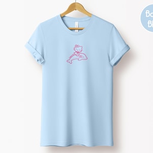 May include: A light blue t-shirt with a pink graphic of a cat character riding a dolphin. The t-shirt is hanging on a wooden hanger against a white background. The words "Baby Blue" are in a blue circle in the upper right corner.