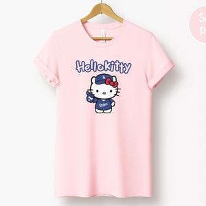 May include: A pale pink t-shirt with a Hello Kitty graphic wearing a baseball cap and jersey with the word "Dodgers". The text "Hello Kitty" is above the graphic in blue. The shirt is hanging on a wooden hanger.