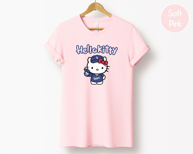 May include: A pale pink t-shirt with a Hello Kitty graphic wearing a baseball cap and jersey with the word "Dodgers". The text "Hello Kitty" is above the graphic in blue. The shirt is hanging on a wooden hanger.