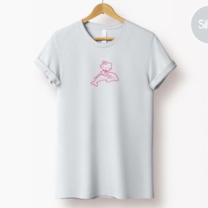 May include: A light gray t-shirt featuring a pink graphic of a cartoon cat riding a dolphin. The cat has a bow and is smiling. The t-shirt is hanging on a wooden hanger.