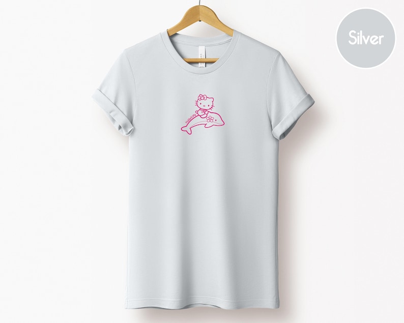 May include: A light gray t-shirt featuring a pink graphic of a cartoon cat riding a dolphin. The cat has a bow and is smiling. The t-shirt is hanging on a wooden hanger.